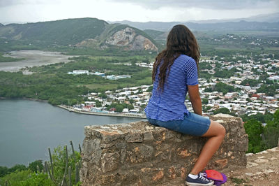 Full length of woman sitting on retaining wall by river against mountains