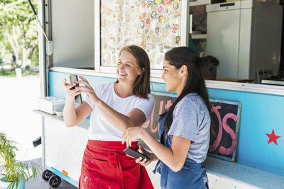 Happy young multi-ethnic female owners using smart phone while standing at concession stand