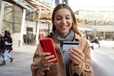 Young casual woman smiling happy holding her smartphone and enters credit card number outdoors