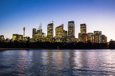 Illuminated buildings by river against clear sky