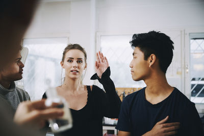 Young female student standing with raised hand amidst males looking at teacher in chemistry laboratory