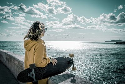 Woman holding skateboard looking at sea against sky