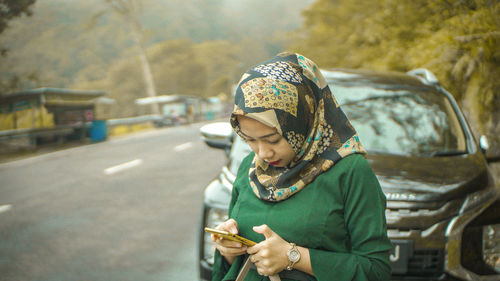 Young woman in hijab using mobile phone by car on road