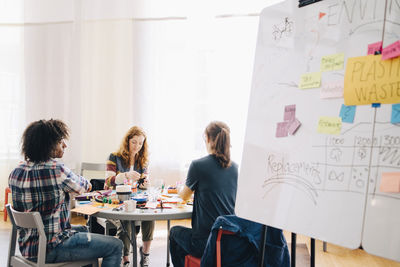 Multi-ethnic business colleagues working at table by whiteboard in creative office
