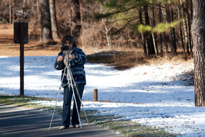 Full length of woman photographing with camera on tripod at forest during winter