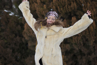 Cheerful young woman with arms outstretched listening music while standing against trees in park during winter