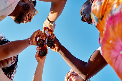 From below of cheerful young diverse best friends smiling and toasting with bottles of beer against cloudless blue sky on sunny summer day