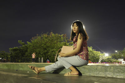 Young woman looking away while sitting on illuminated tree against sky