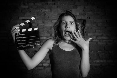 Portrait of surprise woman holding film slate while standing against brick wall