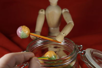 Cropped hand holding lollipop in jar with figurine against red fabric