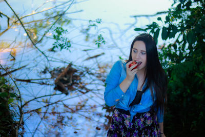 Woman eating apple while standing against trees
