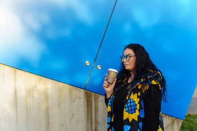 Woman with long curly hair in black, blue and yellow waistcoat drinks coffee from paper cup 