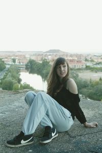 Young woman sitting on concrete wall by river against clear sky