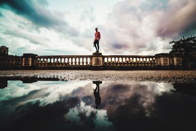 Low angle view of man standing on retaining wall with reflection on pond