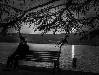 Woman sitting on bench in park by lake against sky