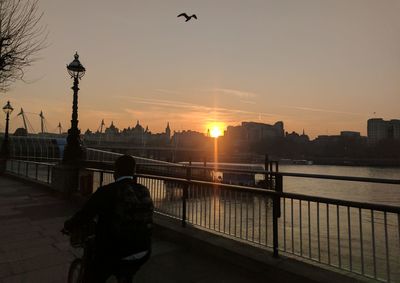 Silhouette man standing on railing by river against sky during sunset