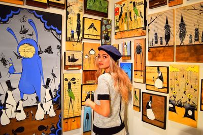 Portrait of young woman standing by various paintings on wall