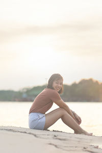 Portrait of young woman sitting on shore at beach against sky