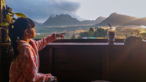 Side view of girl pointing while looking at mountains against sky