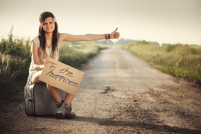 Full length portrait of woman hitchhiking while holding text on road