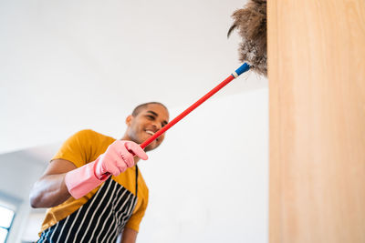 Young man cleaning furniture with broom while standing on stool at home
