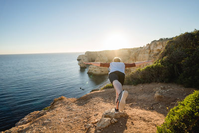 Rear view of playful man on cliff by sea against clear sky
