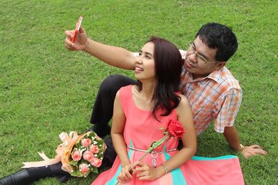 High angle view of smiling couple taking selfie while sitting on grassy field