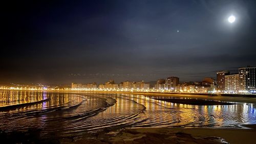 Illuminated city by river against sky at night