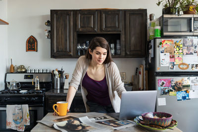 Young woman using laptop while having coffee at table in kitchen