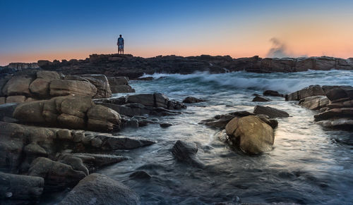 Mid distance view of people standing at coolum beach against sky during sunset