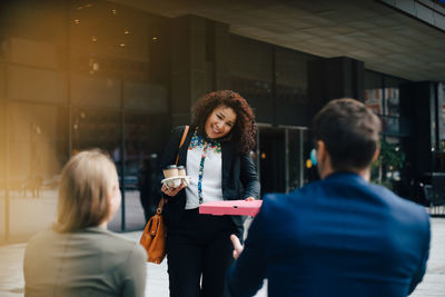 Smiling mid adult businesswoman talking on mobile phone while giving food and drinks to colleagues against cafe