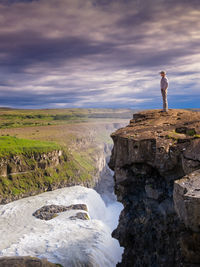 Side view of mature man standing on cliff by waterfall against cloudy sky