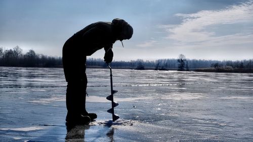 Man drilling in frozen lake against sky during winter