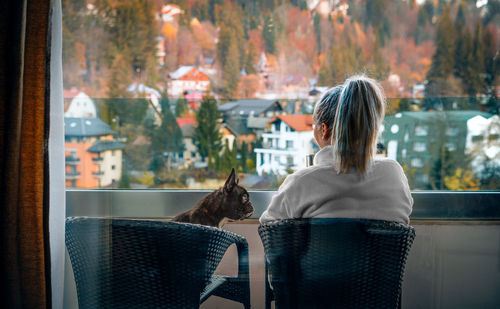 French bulldog and woman drinking morning coffee in balcony against mountain town in autumn