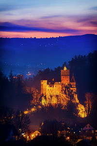 Illuminated bran castle by mountain against sky at dusk