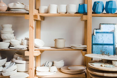 Collection of handmade ceramic bowls and vases with pots and plates with old mirror near different types of utensil standing on wooden shelves in light studio