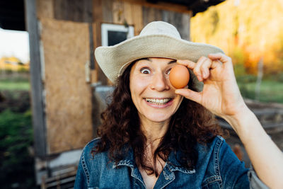 Laughing woman in hat holds egg to face, closing eye