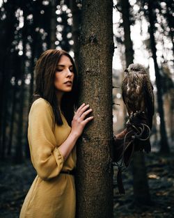 Beautiful woman standing with hawk by tree in forest