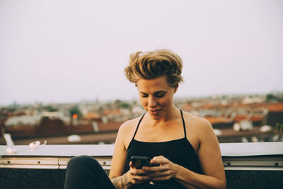 Smiling woman with short hair text messaging on smart phone while sitting on terrace against sky