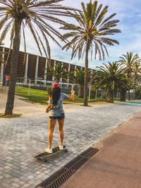 Rear view of woman standing on skateboard against palm trees