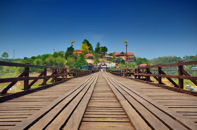 Wooden boardwalk leading towards kanchanaburi against clear blue sky