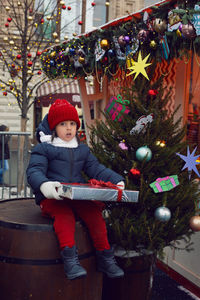 Christmas boy a child in warm clothes holds a gift and sits on a barrel next to a christmas tree