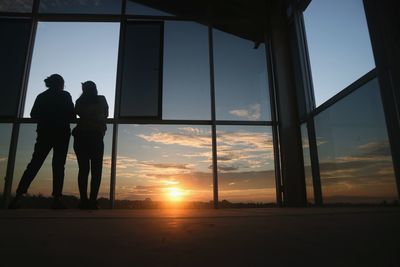 Low angle view silhouette of people standing by window against sky during sunset