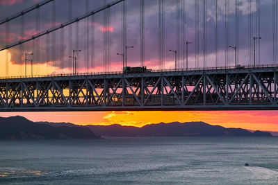 Silhouette bridge over sea against moody sky at sunrise