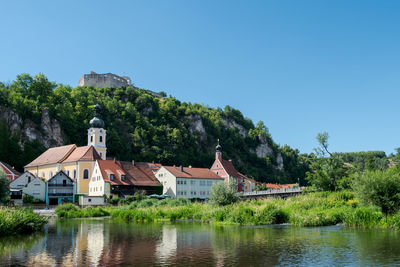 Houses by river and buildings against clear blue sky