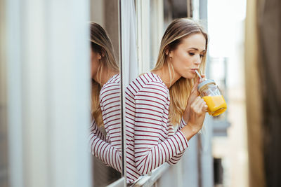 Young woman drinking juice while leaning out of window