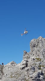 Low angle view of helicopter flying over rocky mountain against clear blue sky