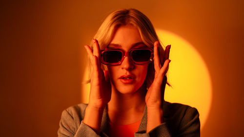 Fashionable model wearing trendy sunglasses and stylish hat against orange background in studio