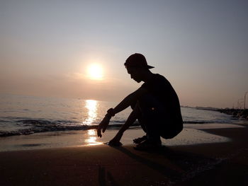 Silhouette man crouching at beach against sky during sunset