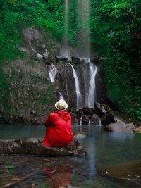 Rear view of man sitting on rock against waterfall in forest